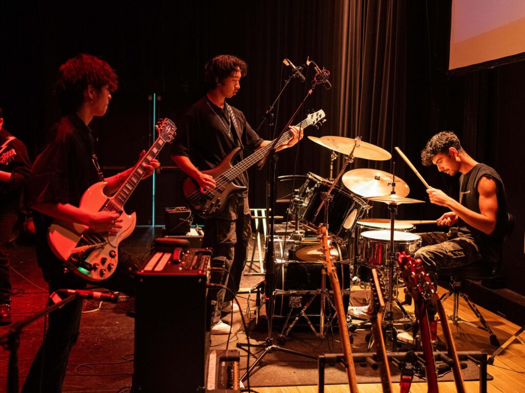 Three musicians playing musical instruments together. Two musicians are playing guitars and another is playing a drumset.